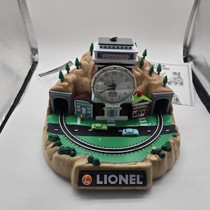 Puede incluir: Un reloj Lionel con una escena de ciudad en miniatura, que incluye una esfera de reloj, edificios, túneles y una vía de tren. El reloj tiene una base marrón con el logotipo de Lionel y un paisaje verde y marrón.
