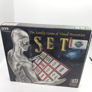 May include: A box for the computer game "SET", a family game of visual perception. The box features a silver robot-like figure holding a game board with symbols. The game is selected by Games Magazine.