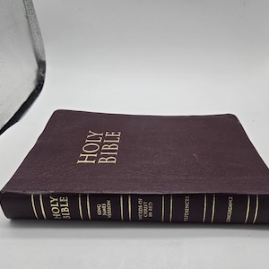 Puede incluir: Una Biblia Sagrada encuadernada en cuero burdeos con letras doradas. El libro está tumbado, mostrando el título "Holy Bible" en la portada. El lomo incluye el texto "King James Version" y otros detalles.
