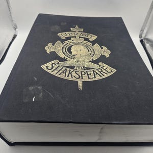 Puede incluir: Un libro negro de tapa dura con letras doradas en la portada. El título es "The Library of Shakespeare".