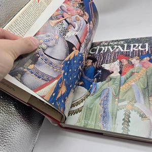 May include: Open book with colorful illustrations. One page shows a knight on a horse, the other displays a group of people in medieval attire. The book's cover reads "The Age of Chivalry" by National Geographic Society.