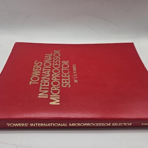 May include: A red book titled "Towers' International Microprocessor Selector" with gold lettering. The book is by T.D. Towers. The title is also printed along the bottom edge. The book is lying on a white surface.