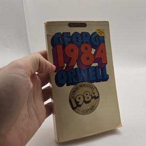 May include: A paperback copy of the book 1984 by George Orwell, Signet Classic edition. The cover features bold blue and red block lettering against a cream background, with a circular Commemorative 1984 Edition seal printed in the center.