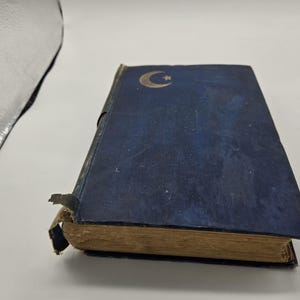 Op de afbeelding: Een vintage boek met een donkerblauwe kaft met een gouden maan en ster ontwerp. De randen van het boek zijn versleten en de rug vertoont tekenen van ouderdom. De pagina's zijn vergeeld, wat duidt op de leeftijd. Het boek staat tegen een witte achtergrond.