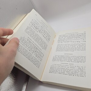 Puede incluir: Un libro abierto con texto y jerogl&iacute;ficos. Las p&aacute;ginas son de color blanquecino con texto negro. El libro est&aacute; abierto por una mano, revelando el texto y las ilustraciones. El lomo del libro es visible.