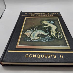 Op de afbeelding: Een zwart hardcover boek met de titel "A Picturesque Tale of Progress" met gouden letters. De omslag toont een ingelijste illustratie van een cherubijn die op een fiets rijdt die door twee vissen wordt getrokken. Het boek heet "Conquests II".