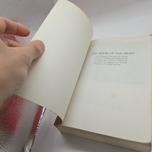 Puede incluir: Un libro abierto titulado "THE BOOK OF THE DEAD" con texto visible en la p&aacute;gina derecha. Una mano est&aacute; pasando la p&aacute;gina de la izquierda. El libro tiene p&aacute;ginas blanquecinas y una cubierta desgastada. El texto incluye el nombre del autor y la afiliaci&oacute;n al Museo Brit&aacute;nico.
