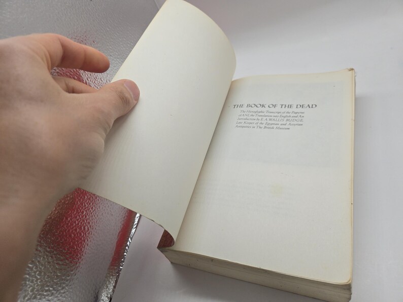 Puede incluir: Un libro abierto titulado "THE BOOK OF THE DEAD" con texto visible en la p&aacute;gina derecha. Una mano est&aacute; pasando la p&aacute;gina de la izquierda. El libro tiene p&aacute;ginas blanquecinas y una cubierta desgastada. El texto incluye el nombre del autor y la afiliaci&oacute;n al Museo Brit&aacute;nico.