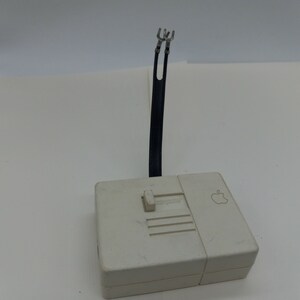May include: White Apple Computer SCSI to DB-9 Serial Adapter with a black cable and two metal prongs. The adapter has a switch labeled "Computer/Peripheral".