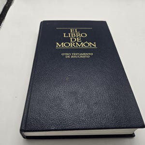 Op de afbeelding: Een marineblauw boek met gouden letters. De titel luidt "EL LIBRO DE MORMON" en "OTRO TESTAMENTO DE JESUCRISTO". Het boek heeft een gestructureerde kaft en witgerande pagina's. Het boek is gesloten en ligt op een wit oppervlak.