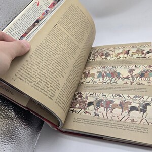 May include: An open book with pages displaying text and illustrations of historical scenes. The book is held open by a hand, revealing detailed drawings of people on horseback and in battle. The page number 104 is visible.