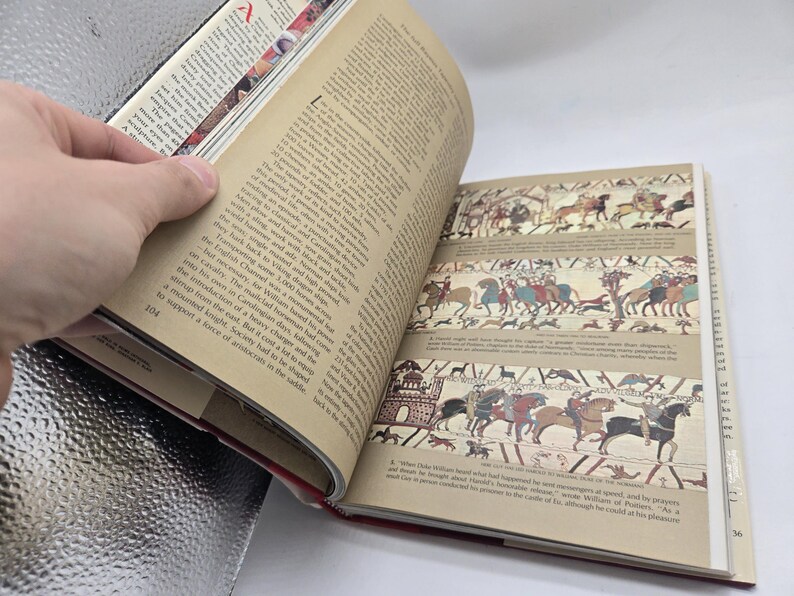 May include: An open book with pages displaying text and illustrations of historical scenes. The book is held open by a hand, revealing detailed drawings of people on horseback and in battle. The page number 104 is visible.