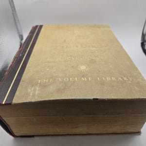 May include: An old book with a tan cover and gold lettering. The book's title, "The Volume Library," is visible, along with the word "encyclopedia." The spine is dark brown with gold stripes. The pages are aged, and the book appears well-used.