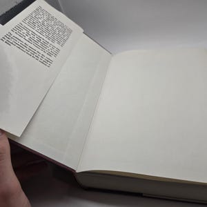 May include: A closed hardcover book with a white cover and a red spine. The book has a glossy cover and a small amount of text on the spine.