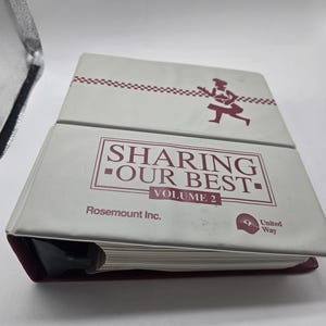 Puede incluir: Una carpeta blanca con el texto "SHARING OUR BEST VOLUME 2" en color granate. La carpeta presenta un gráfico de chef y el logotipo de United Way. La carpeta es de Rosemount Inc.