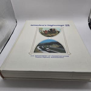 以下が含まれることがあります： 「America's Highways 1776-1976」というタイトルのハードカバー本。白い表紙で、砂時計のデザインに馬車と現代の高速道路のイラストが描かれています。下部には米国運輸省のロゴがあります。