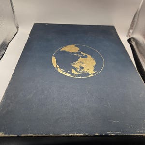 May include: A rectangular, dark blue book with a gold-colored world map emblem. The book's cover shows signs of wear, with a slightly distressed appearance. The emblem is a detailed depiction of the Earth.
