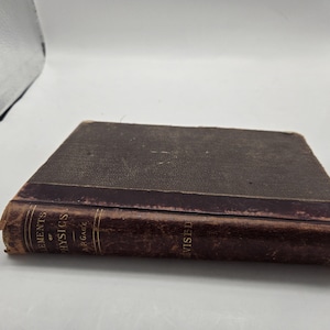 Puede incluir: Un libro antiguo con una cubierta marrón oscuro y desgastada. El lomo presenta letras doradas que dicen "ELEMENTS OF PHYSICS" y "REVISED". Los bordes del libro muestran signos de edad y desgaste, lo que sugiere que es un artículo vintage.