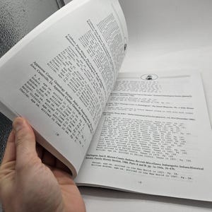 May include: An open book with white pages, featuring printed text. The pages are being turned, revealing more text. The text includes names, ages, and locations. The book appears to be a genealogy or historical reference.