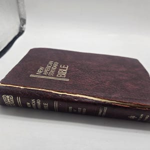 May include: A maroon leather-bound book titled "New American Standard Bible." The book has gold lettering and decorative accents on the spine. The edges of the pages are yellowed, and the cover shows signs of wear.