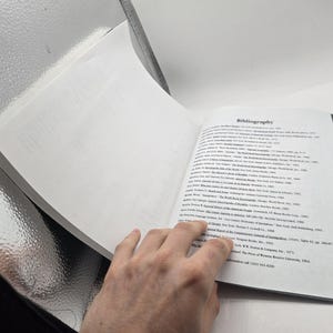 May include: Open book with the title "Bibliography" on the right page. The left page is blank. The book is open, revealing text. The book is held by a hand.
