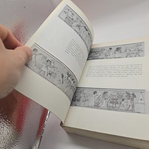 Puede incluir: Un libro abierto con p&aacute;ginas color crema que muestran ilustraciones en blanco y negro de arte egipcio antiguo. Una mano sostiene el libro abierto. Las ilustraciones representan figuras y escenas de la vida egipcia antigua.
