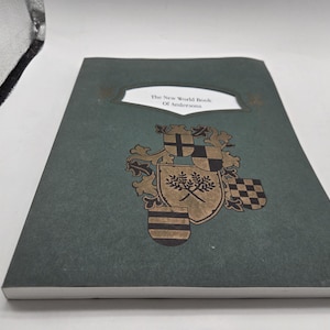 May include: A hardcover book with a dark green cover. The title "The New World Book Of Andersen" is displayed in a white rectangular frame. A gold and black heraldic crest is featured below the title, adding a vintage aesthetic.