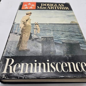 Op de afbeelding: Een zwart hardback boek met een witte titel "Reminiscences" en de naam van de auteur "Douglas MacArthur" in wit op de voorkant. De omslag toont een zwart-witfoto van een man in militair uniform die op het dek van een schip staat.