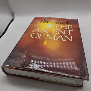 以下が含まれることがあります： J.ブロノフスキー著のハードカバー本「The Ascent of Man」。表紙はオレンジと茶色のグラデーションになっています。ブックカバーには使用感があり、下端に目に見えるシワや剥がれがあります。本は斜めに置かれています。