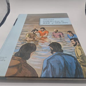 Könnte beinhalten: Ein Hardcover-Buch mit dem Titel "Illustrierte Geschichten aus dem Buch Mormon". Das Cover zeigt eine Illustration von Menschen im Wasser, mit Figuren in verschiedenfarbigen Gewändern. Das Buch hat einen hellblauen Buchrücken.