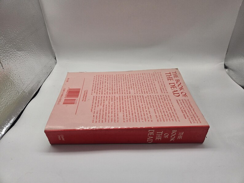 Puede incluir: Un libro de tapa dura titulado "The Book of the Dead" con lomo rojo y cubierta rosa claro. El t&iacute;tulo del libro est&aacute; impreso verticalmente en el lomo. La portada presenta texto impreso y un c&oacute;digo de barras.