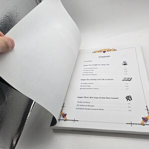 May include: Open book with a white cover, displaying the contents page. The page features a decorative border with swords and crests. The hand is turning a page, revealing the blank side.