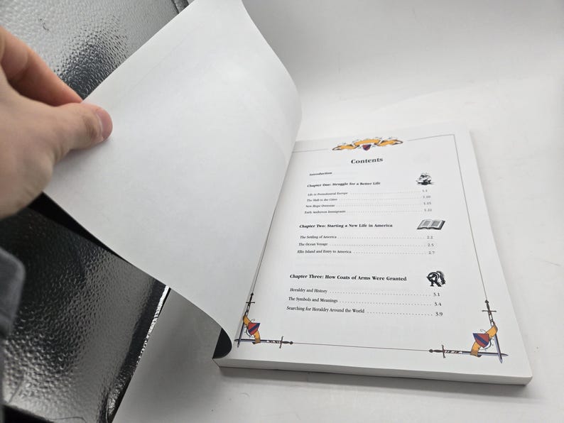 May include: Open book with a white cover, displaying the contents page. The page features a decorative border with swords and crests. The hand is turning a page, revealing the blank side.