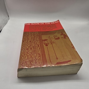 Puede incluir: Un libro antiguo titulado "THE BOOK OF THE DEAD" con una cubierta roja y marr&oacute;n. La cubierta presenta jerogl&iacute;ficos e ilustraciones en marr&oacute;n y rojo. Las p&aacute;ginas del libro est&aacute;n envejecidas y el lomo muestra desgaste.
