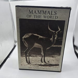 Puede incluir: Un libro titulado "Mamíferos del mundo, segunda edición" presenta una imagen en tonos sepia de un antílope con grandes cuernos. El libro es rectangular y se alza sobre un fondo blanco.