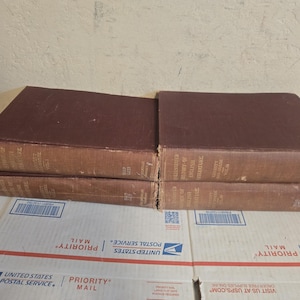 Puede incluir: Cuatro libros antiguos con tapas marrones están apilados. Los libros tienen letras doradas en los lomos y descansan sobre una superficie blanca con texto rojo y azul. Los libros parecen ser antiguos.