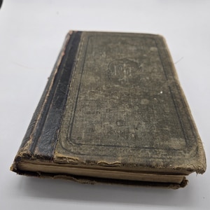 Puede incluir: Un libro antiguo con una cubierta desgastada y texturizada en tonos verdes y marrones. El lomo es negro y los bordes de las páginas muestran signos de desgaste. La cubierta presenta un diseño y texto descoloridos, lo que sugiere que es un artículo vintage.