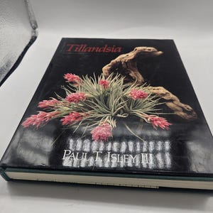 May include: A hardcover book titled Tillandsia by Paul T. Isley III. The black cover features a photograph of a Tillandsia air plant with pink flowers and green foliage attached to a piece of driftwood. The title is in red and the author name is in white.