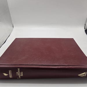 Puede incluir: Un libro de tapa dura de color burdeos titulado "TouchPoint Bible" con letras doradas. El libro está cerrado y tumbado, con el lomo hacia arriba. El logotipo de New Living Translation es visible.