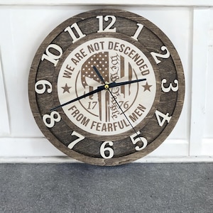 May include: A round wooden wall clock with a dark brown frame and a light brown center. The clock face features the text "WE ARE NOT DESCENDED FROM FEARFUL MEN" and a skull design. The clock hands are black.