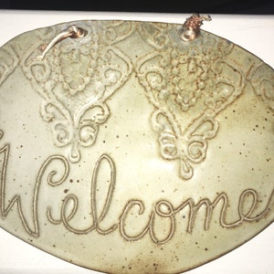 May include: Oval ceramic welcome sign with a light green glaze and brown speckles. The word "Welcome" is written in cursive script. The sign has a decorative floral pattern and a copper wire for hanging.