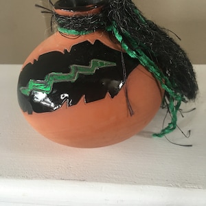 May include: A small, terracotta pot with a black and green design. The pot has a black glaze with a green lightning bolt design. The pot has a black tassel hanging from the top.