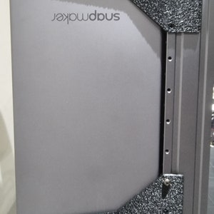 May include: A close-up of a gray, rectangular, metal panel with the word "snapmaker" printed in black. The panel is attached to a black and gray metal frame.