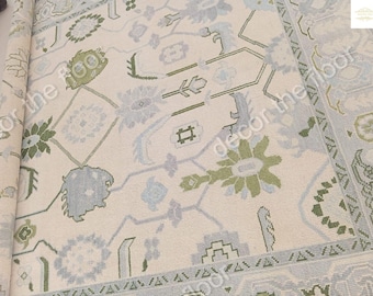 Puede incluir: Una alfombra de color crema con un patrón geométrico en tonos de verde y azul. La alfombra tiene un brillo sutil y una textura suave.