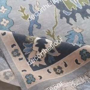 Op de afbeelding: Een blauw en crèmekleurig tapijt met een franje rand. Het tapijt heeft een bloemmotief met een subtiel geometrisch patroon.