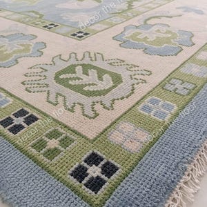 Pode incluir: Close-up de um tapete azul e verde estampado com franjas brancas. O tapete tem um design geométrico com um motivo central de uma flor estilizada.