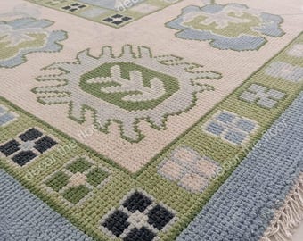 May include: Close-up of a blue and green patterned rug with a white fringe. The rug has a geometric design with a central motif of a stylized flower.