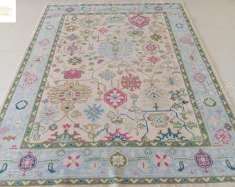 May include: A rectangular rug with a light cream background and a detailed pattern in shades of pink, blue, green, and beige. The border is light blue with floral and geometric designs. The rug is likely made of woven material.