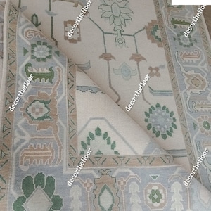 Puede incluir: Una alfombra beige con un patr&oacute;n geom&eacute;trico azul claro y verde. La alfombra tiene una esquina doblada, revelando la parte inferior de la alfombra.