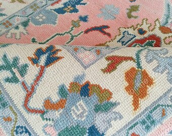 May include: Close-up of two intricately patterned rugs. The top rug is primarily pink with blue, orange, and green floral designs. The bottom rug is rolled up, revealing a white background with similar floral patterns and a pink border.